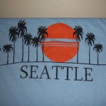Light blue custom t-shirt featuring black palm trees, a large orange sun, and the word "SEATTLE" in bold black letters beneath the graphic—perfect for fans of unique printed tshirts. Rain City West