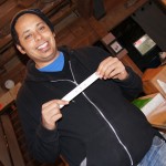 A person in a black hoodie and blue shirt smiles while holding a white strip of paper in a cluttered shop, making you guess who they might be. Rain City West