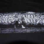 Black t-shirt with a rectangular graphic created using screen printing, featuring a black-and-white photo of graffiti art reading "KRAMS" on a wall. Rain City West