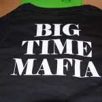 Black T-shirt laid flat on a cardboard box, featuring "BIG TIME MAFIA" in large white letters on the back, crafted with high-quality screen printing. Rain City West