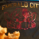 A screen printed t-shirt with "EMERALD CITY" in gold letters and a red geometric design is displayed. A partially eaten donut is visible in the foreground. Rain City West