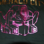 A metallic pink robotic face graphic is screen printed on a dark shirt, with "EMERALD CITY" written in gold letters above it. Rain City West