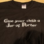 A black t-shirt with white screen printing reads, "Give your child a Jar of Porter," lying flat—perfect for celebrating St. Patrick's Day. Rain City West
