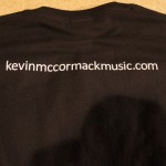 A black t-shirt featuring "kevinmccormackmusic.com" printed in white on the back, crafted with quality screen printing—perfect for St. Patrick's Day celebrations or showing off your musical pride year-round. Rain City West