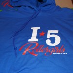 Blue hoodie with "I-5 Rollergirls Seattle, WA" printed on the front in white and red lettering, crafted with quality screen printing for lasting style. Perfect for any screen printing mania fan!. Rain City West