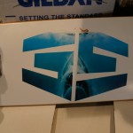 A skateboard from Rain City West with a white deck featuring a stylized blue shark graphic and two metal trucks with white wheels. Rain City West