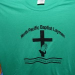Green T-shirt featuring "North Pacific Baptist Layman" text, a cross, and praying hands graphic—perfect to pair with jerseys or hats. Event details below: "Wiletta Faynweather 2012 Walker Gate Layman's Workshop. Rain City West