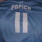 A light blue sports jersey, perfect for fans of custom T-shirts and jerseys, features the name "POPICH" and the number 11 printed in bold white on the back. Rain City West