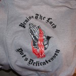 Gray sweatshirt featuring an image of praying hands with a red cloth and the text "Praise The Lord" and "Hats Delicatessen" in black gothic font—a bold piece of deli gear inspired by Dot's Deli. Rain City West