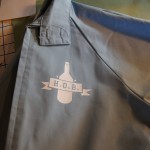 A light blue shirt with a white logo featuring a bottle and a "H.O.B." banner is laid out on a table next to a grid chart and some drawing tools, styled to pair perfectly with vintage jackets. Rain City West