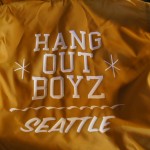 A yellow vintage jacket with white text that reads "HANG OUT BOYZ SEATTLE" printed on the back. Rain City West