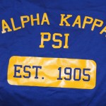 Blue t-shirt with yellow screen printing features "Alpha Kappa Psi" and "EST. 1905" inside a yellow rectangle. Rain City West