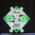 A black t-shirt features a green and white softball-themed screen printing design with crossed bats and text that reads "That's What She Said Softball 2012. Rain City West