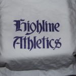 A white t-shirt with the words "Highline Athletics" printed in a dark, gothic-style font. Rain City West