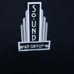 A person holding a black shirt with a white graphic design that reads "SOUND AD GROUP" in stylized text, perfect for showcasing your passion after a day's work. Rain City West