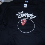 A black T-shirt with “Stones” written in white script and the Rolling Stones tongue logo inside a white circle on the front, crafted with quality by October Screen Printing. Rain City West