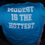 A blue T-shirt with white block letters reads "MODEST IS THE HOTTEST," expertly crafted using Screen Printing, displayed on a pile of other colored shirts. Rain City West