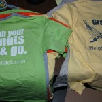 Stacks of T-shirts, screen printed by October Screen Printing, with the text "Grab your nuts & go. WeighMyRack.com" and a graphic of climbing nuts on the back. Rain City West