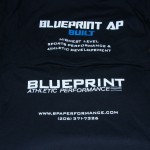 Two black t-shirts with "BLUEPRINT ATHLETIC PERFORMANCE" logos and contact information, screen printed in white and blue text by October Screen Printing, are laid out on a wooden surface. Rain City West