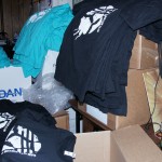 Stacks of folded black and blue t-shirts are arranged on boxes in a cluttered room, with a beer bottle and various items on the table nearby—perfect gear for any Rain City Soccer Club supporter. Rain City West