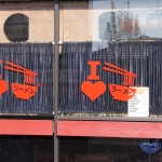 Restaurant window with red graphic of chopsticks holding noodles, a heart symbol, and Japanese text reading "ramen" in orange letters on black curtains. Rain City West