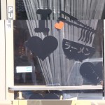 A glass door with drawn black hearts, a bowl with chopsticks, and a red heart. There is a "PULL" sign, a bell, curtains, and reflections visible. Rain City West