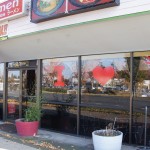 Storefront of a ramen restaurant with large windows displaying "I ♥︎ ramen" graphics, potted plants on the sidewalk, and a red vertical ramen banner. Rain City West