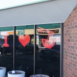 A storefront window displays red graphics of the letter "I," a heart, and a bowl of ramen with Japanese text, with reflections of parked cars visible. Rain City West