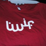 A red TWIF T-shirt with white stylized Arabic script printed on the front, laid flat in a laundry basket next to a blue garment. Rain City West