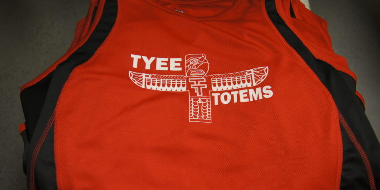 A red athletic sports jersey with black side panels features a white totem pole graphic and the words "TYEE TOTEMS" on the front. Rain City West