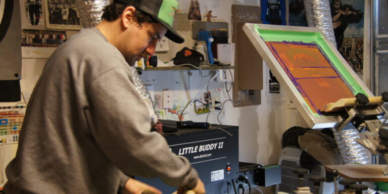 Person wearing a gray sweatshirt and cap screen printing a design onto a shirt in a workshop filled with equipment and posters, utilizing in-store analytics to enhance shop visitor tracking. Rain City West