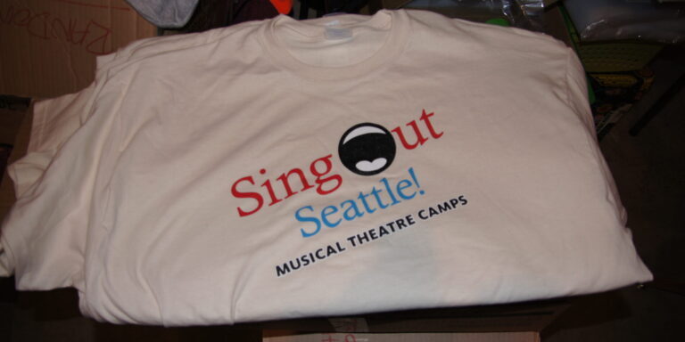 A folded white Sing Out Seattle T-shirt features "Sing Out Seattle! Musical Theatre Camps" and a graphic of an open mouth printed on the front. Rain City West