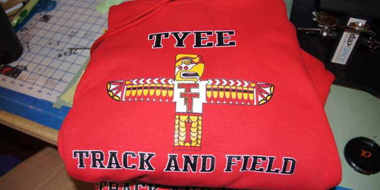 A red Tyee Track & Field sweatshirt with a totem pole graphic and bold lettering, perfect for pairing with your favorite tees and hoodies, lies on a cluttered table. Rain City West