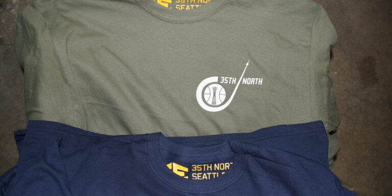Two folded t-shirts, one olive green and one navy blue, with "35th North Seattle" logos printed on the chest and inside neck area—perfect for those who love what we do. Rain City West