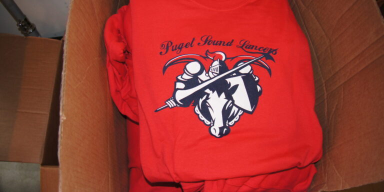 A cardboard box filled with folded red T-shirts featuring a "Puget Sound Lancers" logo with a horse and lance graphic. Rain City West
