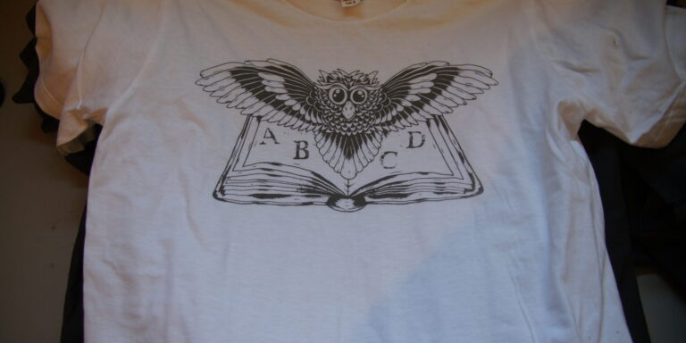 White T-shirt featuring a black line drawing of an open book with an owl above it, wings spread, and the letters A, B, C, and D on the book's pages. Rain City West