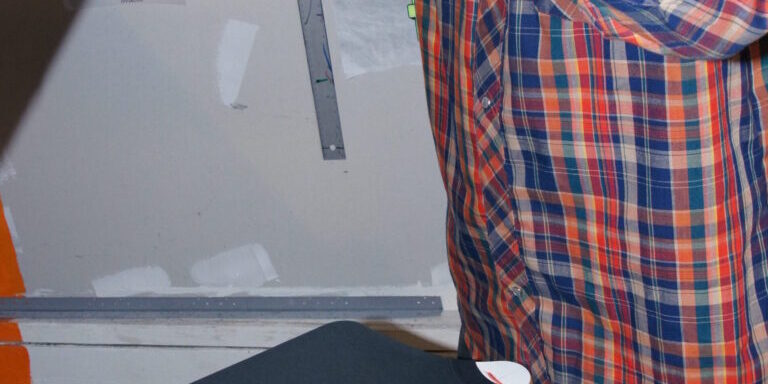Rick, wearing a plaid shirt, uses a heat press in his workspace to print a "Katsu Burger" graphic on black t-shirts. Rain City West