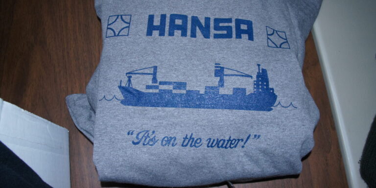 Gray t-shirt with a blue graphic of a cargo ship, the word "HANSA," two logos, and the phrase "It's On The Water!"—a creation inspired by Kyle—printed on the front. Rain City West