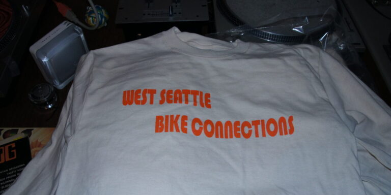 A white shirt with "West Seattle Bike Connections" printed in bold orange letters lies on a dark surface, perfect for fans of local bike connections and stylish shirts. Rain City West
