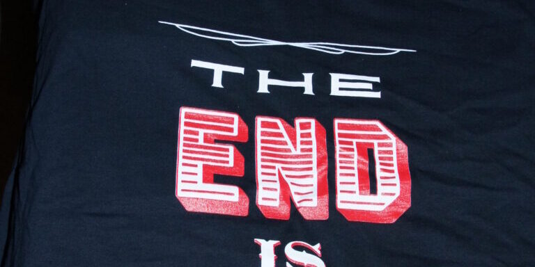 A black sausage-themed t-shirt with “THE END IS HERE” and “SAUSAGE SKATEBOARDS” in bold white and red lettering, displayed on a table alongside other folded shirts. Rain City West